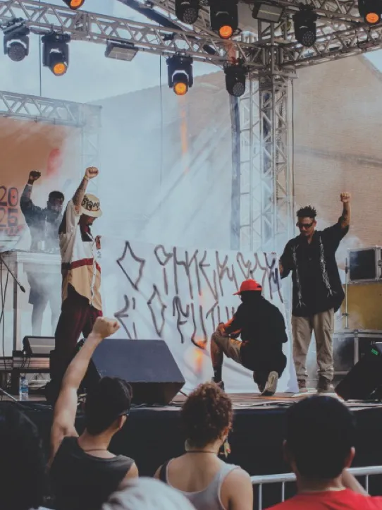 Artistas com o punho erguido no palco do Festival Curau, plateia em primeiro plano